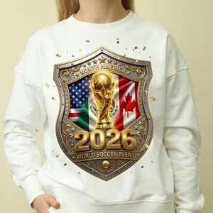 May include: A white long-sleeve sweatshirt featuring a central graphic of a gold trophy inside a shield. The shield displays the flags of the United States, Mexico, and Canada, with the text NORTH AMERICA, 2026, and WORLD SOCCER EVENT.