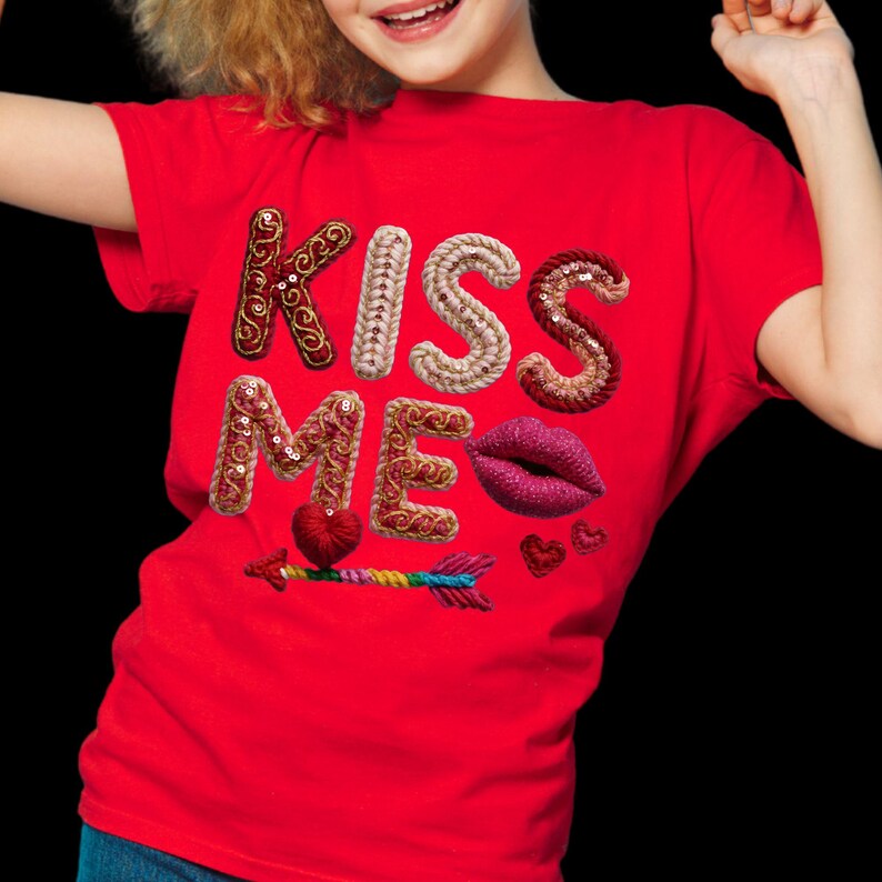 May include: Red t-shirt with the words "KISS ME" embroidered in various textures and colors. The design includes a heart, lips, and an arrow, creating a Valentine's Day theme. The shirt is a vibrant red.