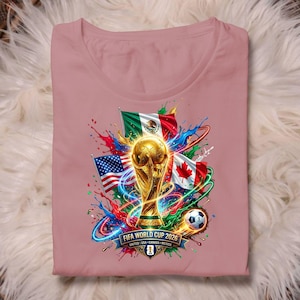 May include: A dusty rose t-shirt featuring a graphic of the FIFA World Cup 2026 trophy, the flags of Mexico, the USA, and Canada, and a soccer ball. The design includes colorful paint splashes and the text FIFA WORLD CUP 2026 UNITED USA CANADA MEXICO.