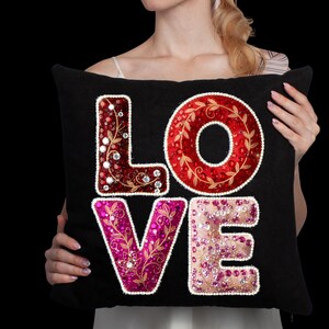 May include: Black decorative pillow with the word "LOVE" in large, embellished letters. The "L" is red with clear gems, the "O" is red with a gold leaf design, the "V" is pink with gold floral accents, and the "E" is pink with clear gems.
