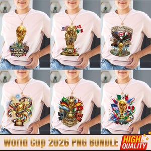 May include: A light pink t-shirt featuring six distinct graphic designs related to the 2026 World Cup. The prints include the golden trophy, soccer balls, national flags of North American countries, an eagle, and text reading North America 2026.