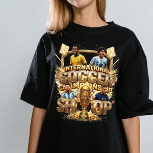 May include: A black t-shirt featuring a graphic print of four soccer players, a golden trophy, and the text "INTERNATIONAL SOCCER CHAMPIONSHIP". The design includes stadium lights, gold accents, and confetti, showcasing players in yellow, blue, and white jerseys.