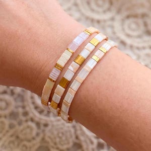 May include: Three beaded bracelets in shades of white, cream, and gold are stacked on a wrist. The bracelets feature rectangular beads with gold accents. The bracelets are a delicate accessory.