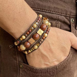May include: Wrap bracelet with a mix of brown, gold, and beige beads. The bracelet has multiple strands with various sizes and textures of beads, including round and square shapes. The bracelet is wrapped around a wrist, showcasing the earthy tones and intricate design.