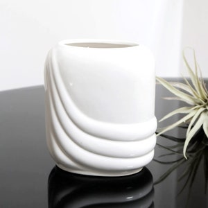 May include: White ceramic vase with a wavy, abstract design. The vase is sitting on a black surface.