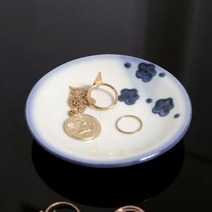 以下が含まれることがあります： 青と白のフラワーデザインが施された白いセラミック製の小物入れ。中には、コインペンダントのついたゴールドチェーンネックレス、ゴールドリング2つ、三角形のデザインのゴールドリングが入っています。
