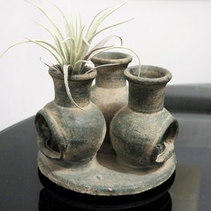 May include: Three small, dark green ceramic pots with a single air plant in the center. The pots are connected to a larger, round base.