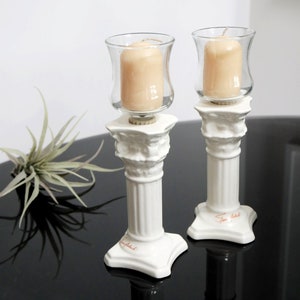 May include: Two white ceramic candle holders with a fluted column design. Each holder has a glass candle cup and a clear glass candle inside. The candle holders are on a black surface.