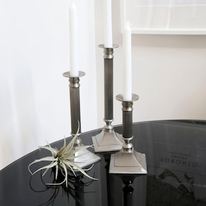 May include: Three silver candle holders with white candles on a black table. The candle holders have a square base and a fluted column. A small green plant is in the foreground.