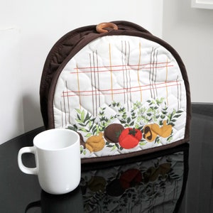 May include: A brown and white quilted coffee pot cover with a wooden handle. The cover has a checkered pattern and a design of vegetables and herbs.