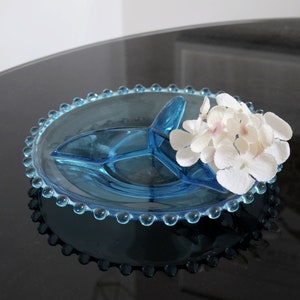 May include: A blue glass divided serving dish with a beaded edge. The dish is on a black surface with white flowers on top.