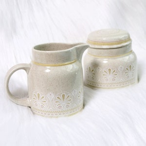 May include: A cream-coloured ceramic creamer and sugar set with a floral design. The creamer has a handle and a spout. The sugar bowl has a lid.