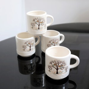 May include: Set of four white ceramic mugs with a brown tree and birds design. The mugs are stacked on top of each other on a black surface.