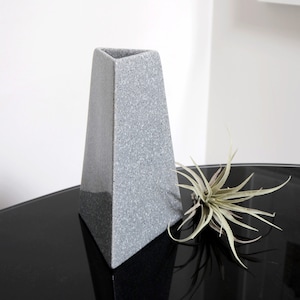 May include: A grey triangular ceramic vase with a speckled texture. The vase is sitting on a black surface with a small green plant.