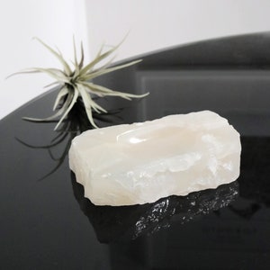 May include: A white, natural, rough-hewn, rectangular, crystal dish with a shallow, rounded depression in the center. The dish is sitting on a black surface.