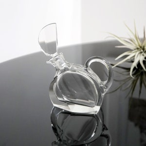 May include: A clear glass decanter with a unique design and a hinged lid. The decanter has a round body and a curved handle.