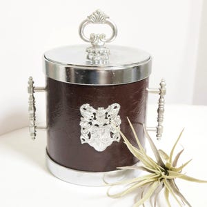 May include: A vintage ice bucket with a brown leather-like exterior and silver-toned accents. The bucket features a decorative handle, a silver lid, and a silver emblem. The bucket is approximately 7 inches tall and 6 inches in diameter.