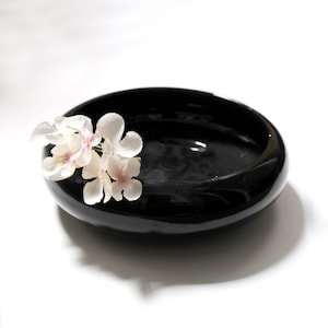 May include: A black ceramic bowl with a white flower arrangement on the rim. The bowl is round and has a glossy finish.