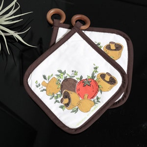 Könnte beinhalten: Ein Paar Topflappen in Braun und Weiß mit einem Pilz- und Tomatenmotiv. Die Topflappen haben einen braunen Rand und einen Schlaufe.