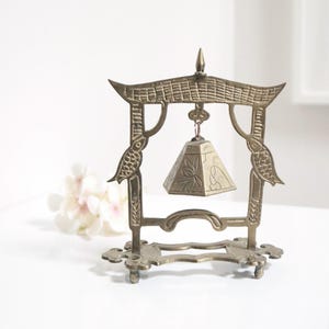 May include: A small, gold-coloured metal bell hanging from a decorative stand. The stand is shaped like a pagoda with intricate details and two birds on either side. The bell is pyramid-shaped with an engraved design.
