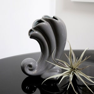 May include: A gray ceramic vase with a unique, wavy design. The vase is shaped like a stylized wave or a seashell. It is sitting on a black surface with a green plant in front of it.