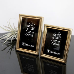 May include: Two gold-toned picture frames with black mats. The frames have a raised edge and are inscribed with the text "Solid Brass Laiton Solide" and "IIC Intercraft".