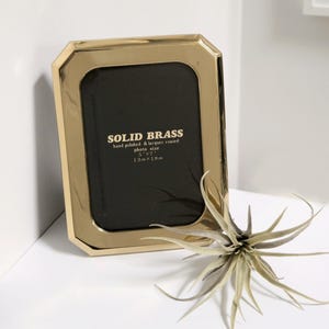 May include: A gold-colored picture frame with a beveled edge and a black mat. The frame is made of solid brass and is hand polished and lacquered. The frame is designed to hold a 5x7 inch photo. The text "SOLID BRASS" is printed on the frame, along with the photo size in inches and centimeters.