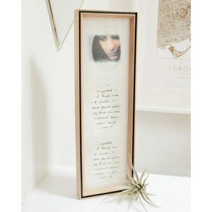 May include: A framed artwork with a light wood frame and a black inner border. The artwork includes a small image of a person's face and handwritten text. A small plant is in front of the frame.