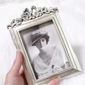 Pu&ograve; includere: Cornice per foto argentata con un designo ornato e una foto in bianco e nero di una donna con un cappello. La cornice misura 3,5 x 5 pollici.