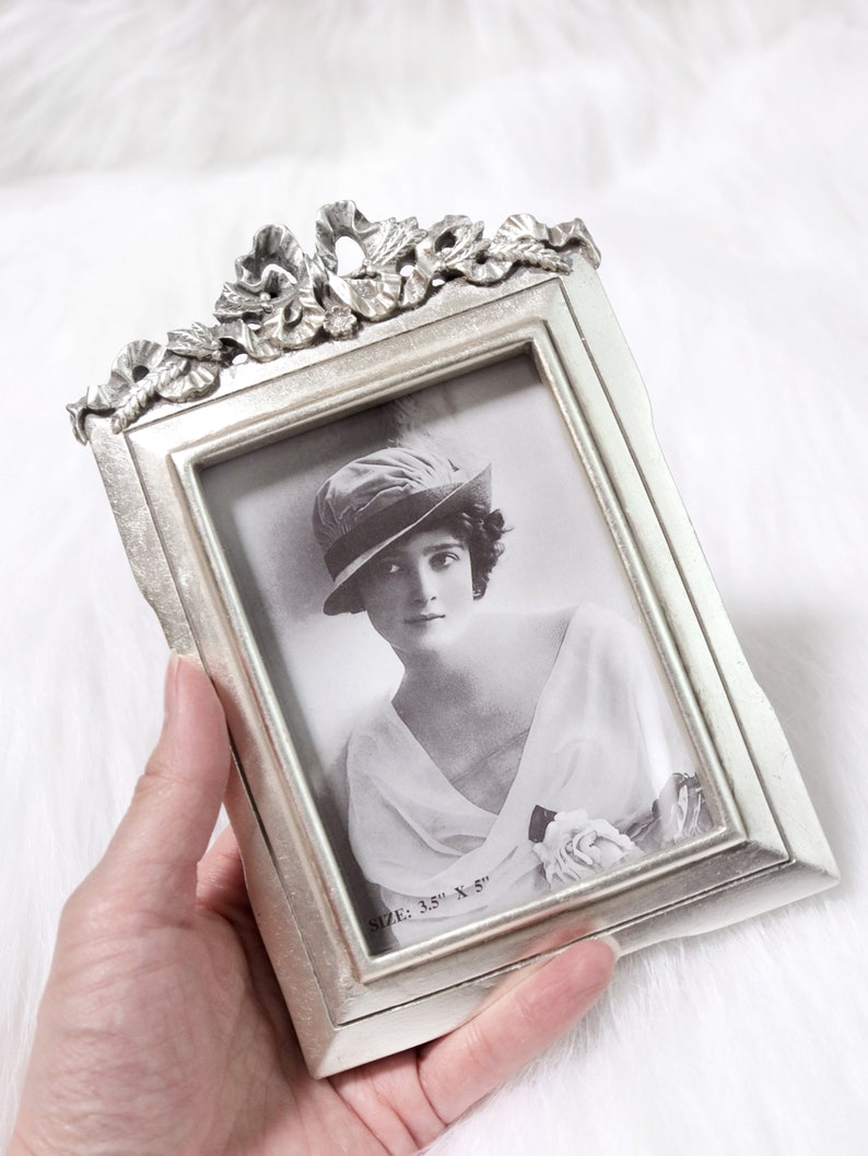 Pu&ograve; includere: Cornice per foto argentata con un designo ornato e una foto in bianco e nero di una donna con un cappello. La cornice misura 3,5 x 5 pollici.