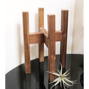 May include: A medium-brown wooden plant stand featuring four vertical legs connected by a central cross-shaped support structure. The stand has a square frame design with a smooth finish, providing a stable base for holding a potted plant.