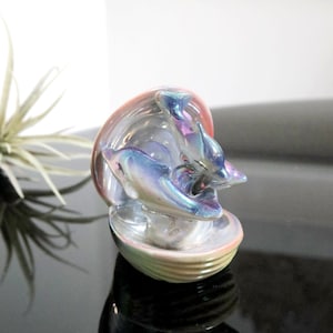 May include: Iridescent glass sculpture of two dolphins leaping from a shell. The sculpture has a pearlescent finish with hues of blue, purple, and pink. The base is a light green color. The sculpture is on a black surface.