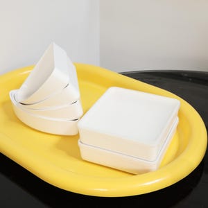 May include: A bright yellow oval tray holds several white, square-shaped containers and a stack of white, triangular-shaped bowls. The containers and bowls are stacked neatly on the tray, creating a clean and organized aesthetic.