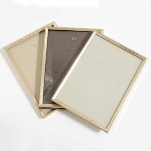 May include: Three rectangular picture frames with gold-coloured metal edges. The frames are arranged at an angle, with one frame showing a light tan backing, another a dark brown backing, and the third a light cream-coloured backing.