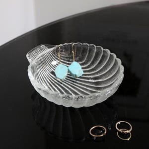 May include: A clear glass seashell-shaped dish with turquoise gemstone earrings inside. The dish is sitting on a black surface.