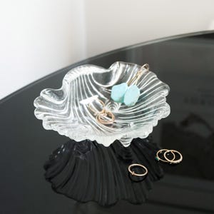 May include: A clear glass dish with a shell-like design. The dish contains small gold rings and turquoise earrings. The dish is on a black surface, with its reflection and the jewellery visible. The dish is likely around 15 cm in diameter.