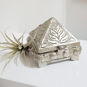 Peut inclure: Une boîte argentée en forme de pyramide avec des motifs complexes de feuilles et de points. La boîte a un couvercle à charnière et repose sur de petits pieds. Un petit bouton transparent sert de fermoir. Une plante aérienne séchée est à côté de la boîte.