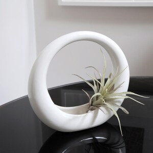 May include: White ceramic planter with a circular opening, designed to hold an air plant. The planter is sitting on a black surface.