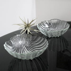 May include: Two clear glass bowls shaped like seashells. The bowls have a textured, ribbed surface and are sitting on a black surface.