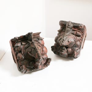 May include: Two antique, hand-carved wooden architectural elements. The pieces are dark brown with reddish-brown accents, featuring detailed relief carvings of stylized faces and floral motifs. The objects are displayed on a white surface.