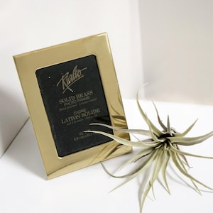 May include: A gold-toned solid brass photo frame with a black mat and the text "Rialto Solid Brass Photo Frame Hand polished - Lacquer coated Cadre Laiton Solide Poli a la main - Laque couché 3 1/2 x 5 8.9 cm x 12.7 cm".
