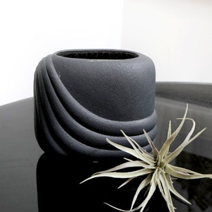 May include: A black ceramic planter with a wavy, textured design. The planter is empty and sitting on a black surface. A small air plant is in the foreground.