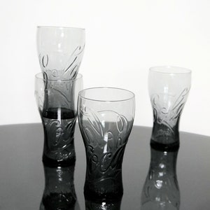Könnte beinhalten: Vier klare Glasbecher mit einem matten, grauen Finish. Die Gläser haben ein Coca-Cola-Logo, das in das Glas geätzt ist.
