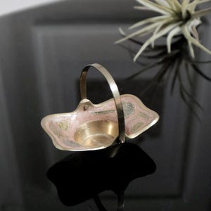 Può includere: Un piccolo cestino decorativo in ottone con manico. Il cestino è dipinto con motivi floreali rosa, verdi e dorati.