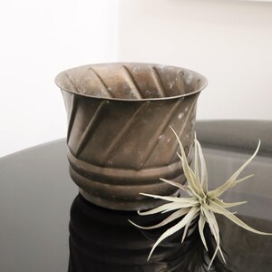 May include: A bronze-colored metal planter with a diagonal ribbed design. The planter has a wide opening and a slightly flared rim. A small, spiky, air plant rests beside the planter, adding a touch of natural texture.