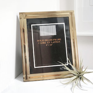 Könnte beinhalten: Ein goldfarbener Bilderrahmen mit schwarzem Passepartout und einem weißen Etikett mit der Aufschrift "SOLID BRASS FRAME CADRE EN LAITON 8" x 10"".