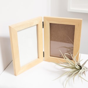 May include: A two-panel wooden picture frame, open and hinged, with a light wood finish. One side has a white backing, the other a brown backing. A small plant with long, thin leaves rests near the frame.