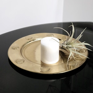 May include: A gold-toned metal tray with a raised shell design around the edge. A white candle sits in the centre of the tray, and a small green plant is placed on the tray to the right of the candle.