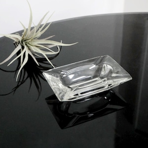 May include: A clear glass ashtray with a rectangular shape. The ashtray has a slightly raised edge and is sitting on a black surface.