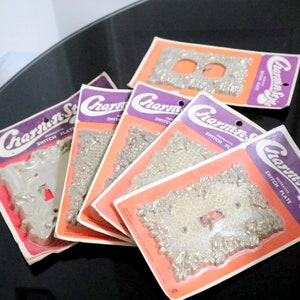 May include: Five packages of decorative switch plates with a floral design. The packages are all white with pink and purple accents. The switch plates are made of a shiny, silver-colored metal. The packages say "Charm-n-Style Decorative Switch Plate" on them.